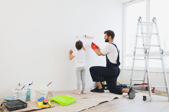 Repair in the apartment. Back view of happy family of father and little cute child daughter paints the wall with white paint.