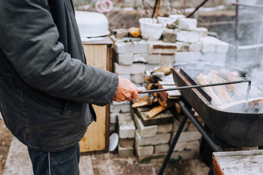 A man in the open air, in nature, holds a metal poker in his hands and corrects burning firewood in the barbecue. Photography, food preparation.