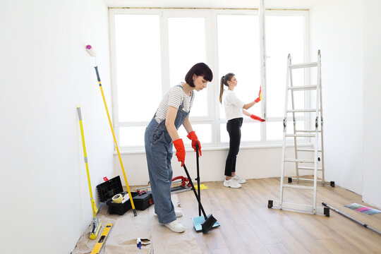 Young beautiful women cleaning up new apartment after renovation. One girl sweeping floors in house with broom and dustpan, and another wiping the window.