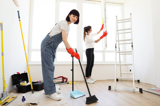 Red haired girl in overalls sweeping floors in house with broom and dustpan, and another female in casual clothes wiping the window. Young beautiful women cleaning up new apartment after renovation.