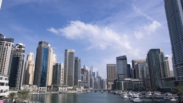 4k, timelapse. View of the towers of Dubai Marina from the bridge to the embankment in the bay on a sunny day. Clouds are floating in the blue sky, yachts are standing and floating, people are walking