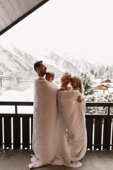 Family standing on balcony covered in blanket in early morning enjoying good winter day in Alps.