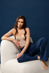 A beautiful stylish girl in blue jeans and a beige corset sits on a chair and looks into the frame. Blue background.