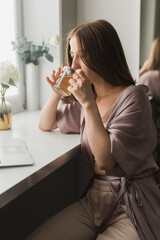 Young woman wears sleeping clothes working on laptop computer while sitting at living room and drinking tea - social network and working at home concept