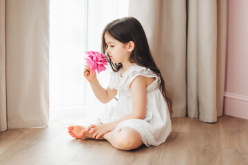 A little girl in a white dress is sitting near the window and holding a rose. A happy child at the...