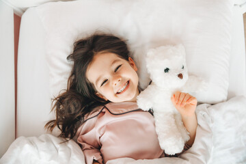 a little dark-haired girl is sleeping in bed, hugging a teddy bear under a white blanket.the child...