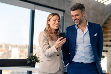 Two successful business persons entrepreneurs man and woman wearing business suits using mobile apps looking at cell phone checking data, smiling working in office.