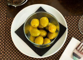 Fresh green olives with pits served on plate with glass and fork nearby