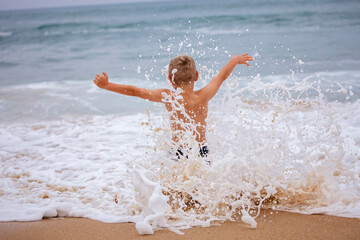 A boy of 7-8 years old sits on the ocean shore with his arms open towards the wind and waves. Storm...