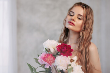 Attractive woman with long blond wavy hair holds her hands near her face. Around her are pink and white fresh flowers in a large beautiful bouquet of flowers. Artistic portrait with flowers.