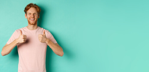 Cheerful bearded man with red hair showing thumbs-up, like and approve something, praising promo, standing against turquoise background