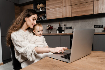 Maternity leave for young mother. Happy family time with daughter at home. Modern childhood. Mom is sitting with toddler child and working on laptop remotely.