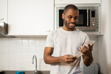 Smiling dark-skinned man reviewing the receipt of the purchase, the offers and the concept of sale, inside his house.