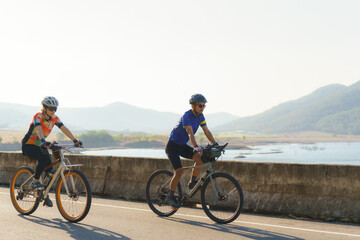 Obraz premium Asian cyclist couple riding together for exercise around the lake in the morning with beautiful mountain view in the background.