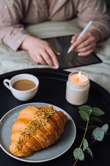 Croissant with cup of cappuccino and scented candle on the tray, woman drawing on digital tablet on background