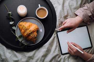 Flat lay, woman using digital tablet for drawing while lying on bed and enjoying croissant with cappuccino