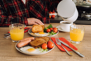 Unrecognizable woman pours water into a cup, prepares to enjoy an English breakfast with coffee and orange juice in the kitchen