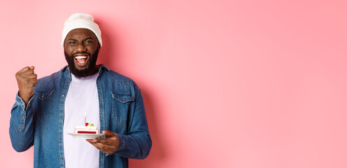 Cheerful african-american guy celebrating birthday, making wish on bday cake with lit candle,...