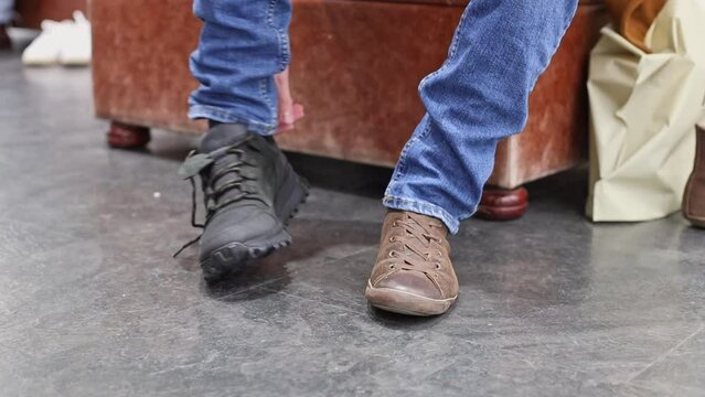 A young handsome caucasian brunette in a brown coat is trying on a dark gray sneaker, touching it with his finger, sitting on a leather pouffe in a shoe store, close-up side view. The concept of offli