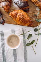 Flat lay of cup of cappuccino with kitchen towel and eucalyptus branch, brown croissants on the cutting board