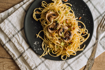 Spaghetti pasta with champignon mushrooms sprinkled with cheese parmesan and green onions on a black plate with a towel and a fork on a wooden table