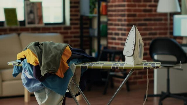 Empty living room with iron and ironing board, large pile of laundered clothes to smooth out creases and wrinkles. Apartment room with appliances and tools to iron garments, house chores.