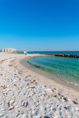 Destin, Florida beach near Noriego Point with seaweeds on white sand shore line. There are rocks on the water on the right and view of modern buildings on the left against the blue sky background.