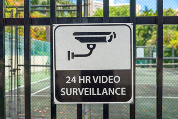 Signage of 24 HR VIDEO SURVEILLANCE on a metal railings of a tennis court in Miami, Florida. Close-up of a sign on a fence railings against the court with mesh screens.