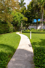Concrete walkway at the entrance of a gated community in Miami, Florida. Narrow walkway in the middle of a green lawn with white lawn lights on the right heading to the white gate railings.