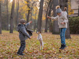 Caucasian children are walking with jack russell terrier in autumn park. Boy, girl and dog are jumping outdoors.