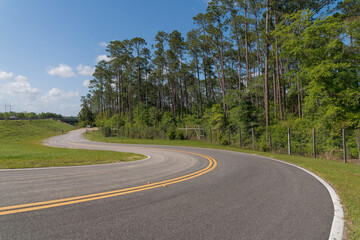 Winding road with double yellow lines in Navarre, Florida. Road near another highway on the left with grass in between and view of a forest trees with metal barriers.