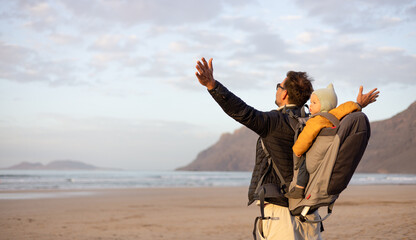 Father rising hands to the sky while enjoying pure nature carrying his infant baby boy son in backpack on windy sandy beach of Famara, Lanzarote island, Spain at sunset. Family travel concept - obrazy, fototapety, plakaty