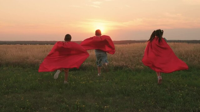 Child hero in red cloak running into sunset. Happy children run, play superheroes against sky. Teamwork, Boy, girl play red cape superhero, childhood dream. Child in raincoat plays. Children nature