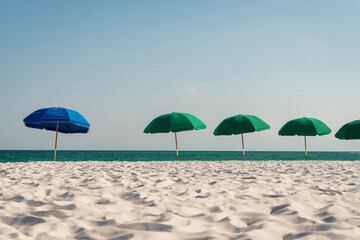Blue beach umbrella among with the green umbrellas in a row at Destin, Florida. White sand on a beach with beach umbrellas against the ocean below clear sky background.