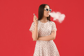 Young woman in sunglasses smoking disposable electronic cigarette on red background