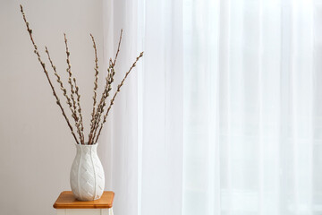 Vase with willow branches on table near light curtain