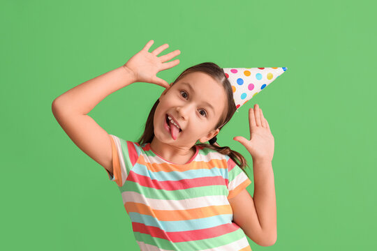 Funny little girl in party hat grimacing on green background. April Fools' Day celebration