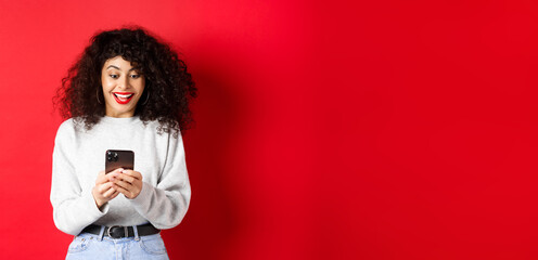 Image of young woman with curly hair, reading message on smartphone and smiling happy, receiving...