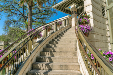 Outdoor stairway going up a building with trees and blue sky background. Beautiful purple flowers hanging on pots decorate the steel railings of the outside stairs.