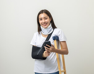 Happy young asian woman broken arm holding smartphone. Woman put on plaster cast splint with walking sticks crutches. Patient wearing sling support arm with neck collar. life insurance and accident - obrazy, fototapety, plakaty