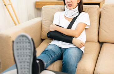 Woman suffered pain from accident fracture broken bone injury with leg splints in cast neck splints collar arm splints sling support arm in living room. Social security and health insurance concept.