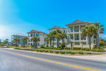 Fototapeta na wymiar Destin, Florida- Fenced beach houses with concrete sidewalks near the highway. There is a highway at the front near the footpath with bushes and palm trees on the side.