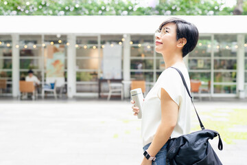 Stylish short hairstyle Asian woman holding, reusable insulated stainless steel water bottle, smile, looking up in front of beautiful cafe in outdoor garden for sustainability living lifestyle