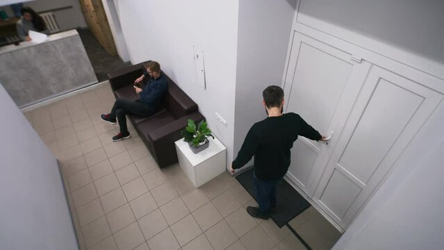 Adult man sits on leather sofa at lobby and waits himself turn. Client comes to reception and talks with female worker. Administrator shows way to cabinet. Business office. Security camera shooting.