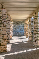 Fototapeta premium Passage with granite path under the roof with rough stone pillars. Vertical shot of a hallway leading to a stone wall at the background.