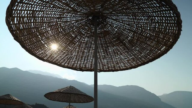 Straw umbrellas on the beach. Blue sky, sea and mountains in the background. Camera movement from top to bottom. The rays of sunlight pass through the straws
