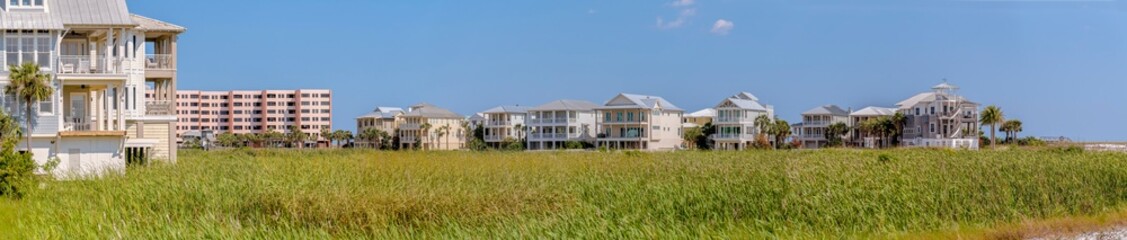 Fototapeta na wymiar Homes on the beach in panorama at Destin point in Destin, Florida. There are tall green grasses at the front of three-storey houses and multi-storey apartment on the left.