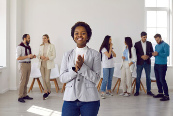 Happy joyful young business lady standing at office on background of her colleagues. African american female team leader or coach standing against background of people communicating with each other.