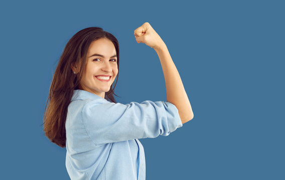 Portrait Of Confident Strong Happy Young Woman Looking At Camera, Smiling Flexing Her Arm Isolated On Blue Background. Girl Power, Strength, Independence Concept. Strong Gesture With Clenching Fist.