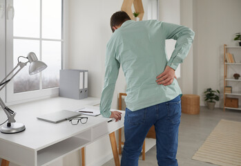 Adult man feels back pain after sedentary work at laptop, sitting at home in uncomfortable position...